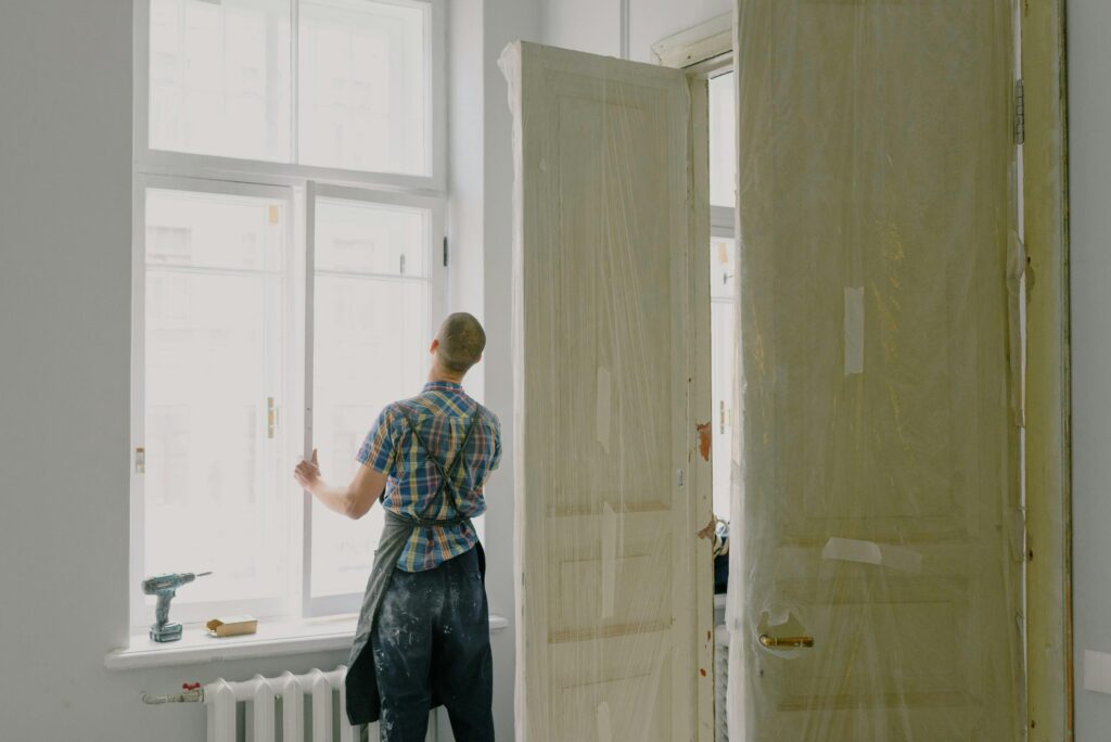 pexels-photo-5691550-5691550 Back view of unrecognizable workman in apron mounting window near drilling machine and doors in flat